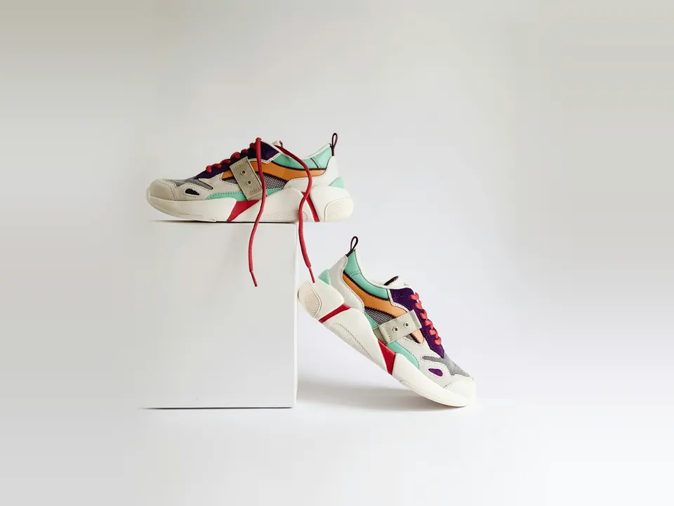 Pair of chunky cream sneakers with mint, orange and purple panels and red laces, one resting on a white box